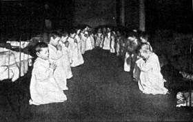Two rows of little boys, about 20 in total, kneel before their beds in ...