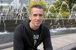 Dan Savage outdoors in a black sweatshirt with a fountain behind him