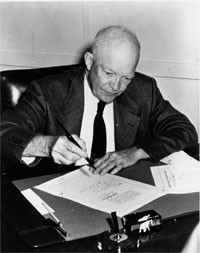 President Eisenhower sits at a desk, signing a piece of paper on top ...