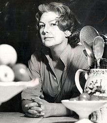 middle aged woman with dark, greying, hair; she is at a kitchen table ...