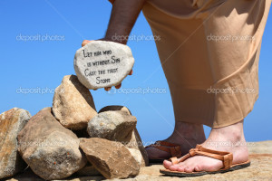 man holds a white rock inscribed with a bible verse from John 8:7 ...