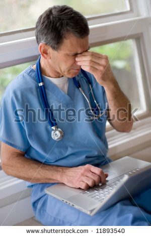 Mature doctor in blue scrubs sitting on floor with laptop, rubbing his ...