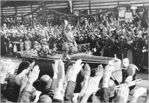 Standing in an open car, Adolf Hitler salutes a crowd lining the ...