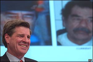 Paul Bremer smiles in front of picture of Saddam Hussein at press